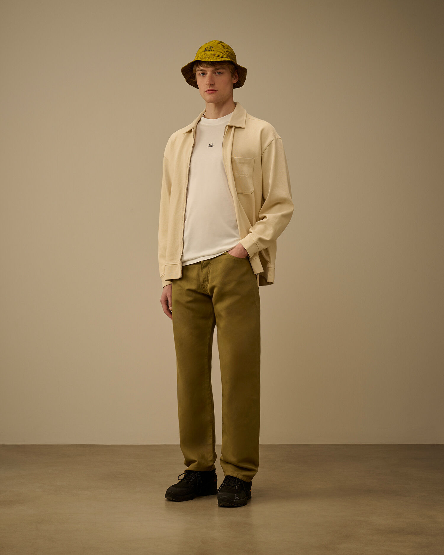 Young man wearing a cream shirt, beige jacket, olive pants, black shoes, and a yellow bucket hat stands against a plain beige background.