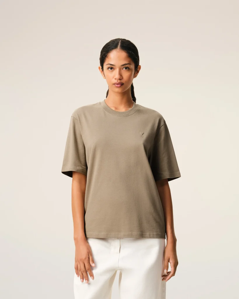 Person wearing an oversized taupe T-shirt and white pants against a neutral beige backdrop, facing the camera.