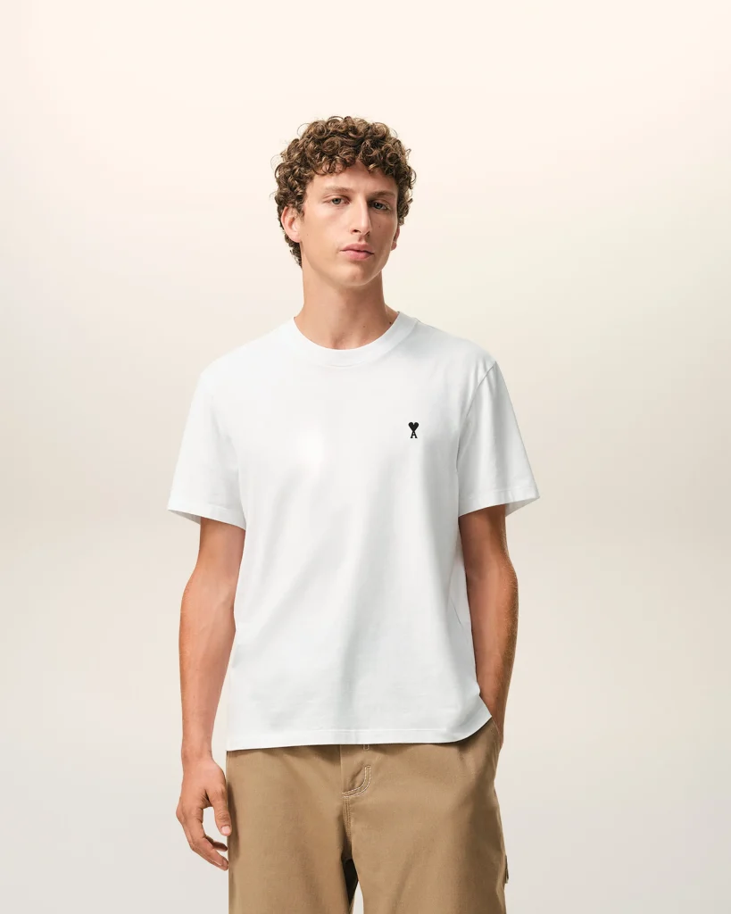 Man in a white T-shirt with a small black chest logo and tan pants, standing against a pale gradient backdrop.