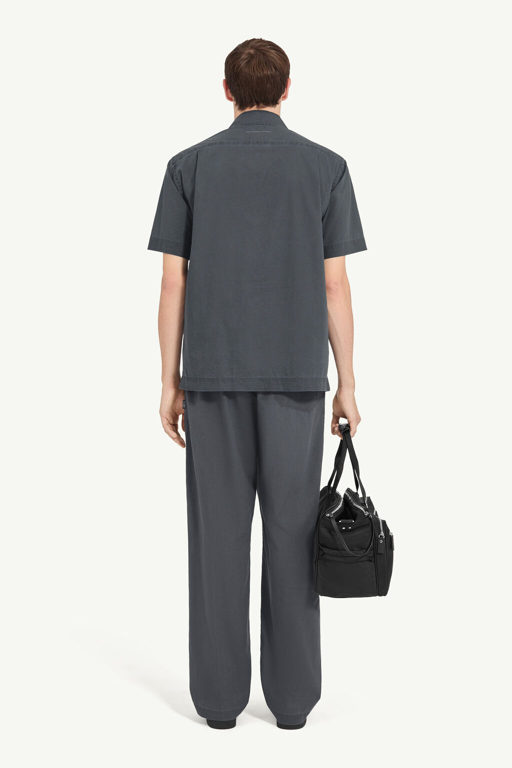 Man seen from behind wearing a loose dark gray short-sleeve shirt and matching wide-leg pants, holding a black duffel bag by the strap.