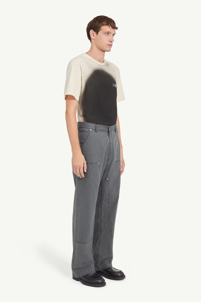 Male model in profile wearing a cream T-shirt with a large black spray-paint design, gray wide-leg cargo pants, and black shoes.