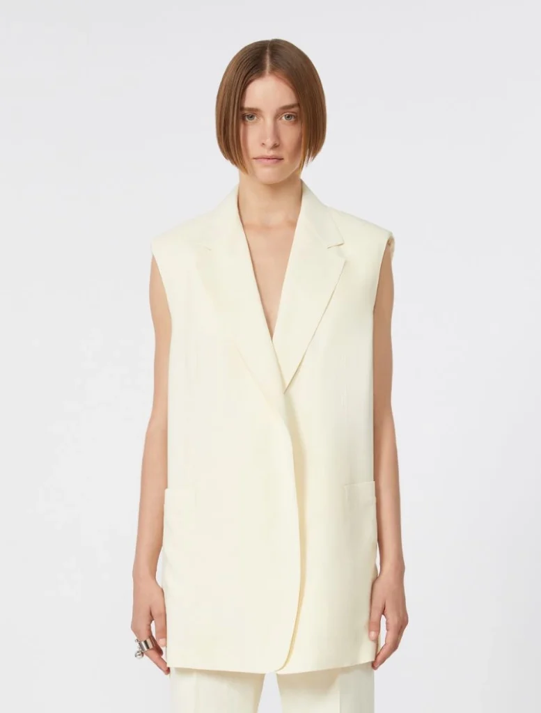 Model wearing a cream sleeveless blazer with wide shoulders and long length over matching pants.