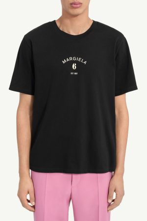 Black crew-neck T-shirt featuring a small white 'MARGIELA 6 EST 1987' logo on the chest, paired with pink trousers on a model.