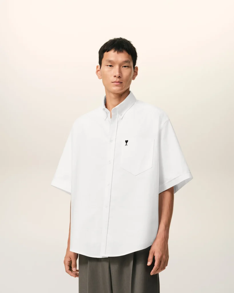 Man in an oversized white button-down shirt with a small black heart on the pocket, standing against a light beige backdrop.