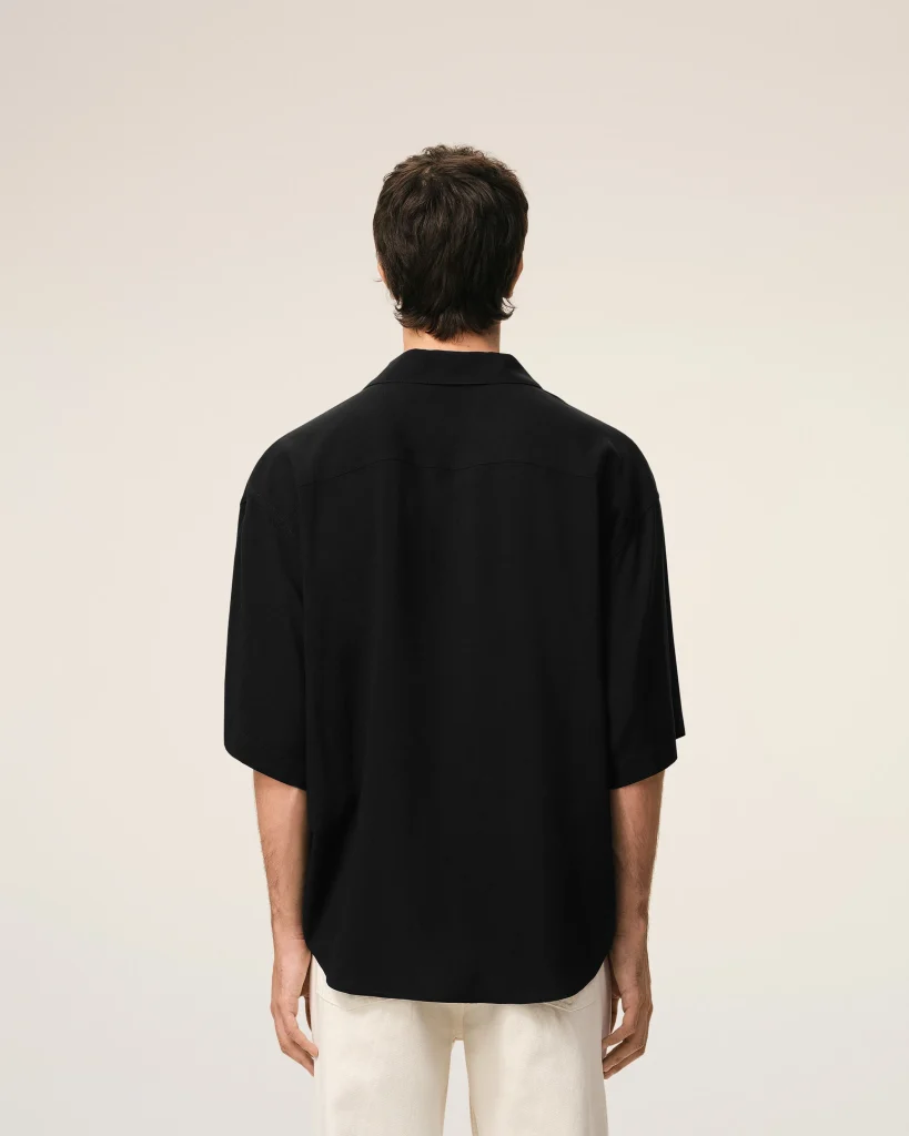 Back view of a person wearing a black shirt, standing against a light neutral background; beige pants are visible at the bottom edge.