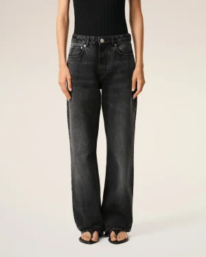 Person from the waist down wearing a black ribbed top tucked into dark washed jeans, standing with open-toe sandals on light background