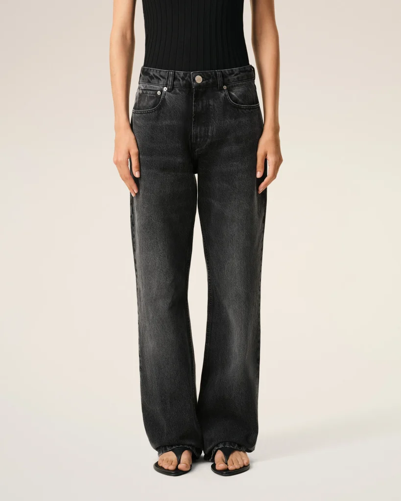 Person from the waist down wearing a black ribbed top tucked into dark washed jeans, standing with open-toe sandals on light background
