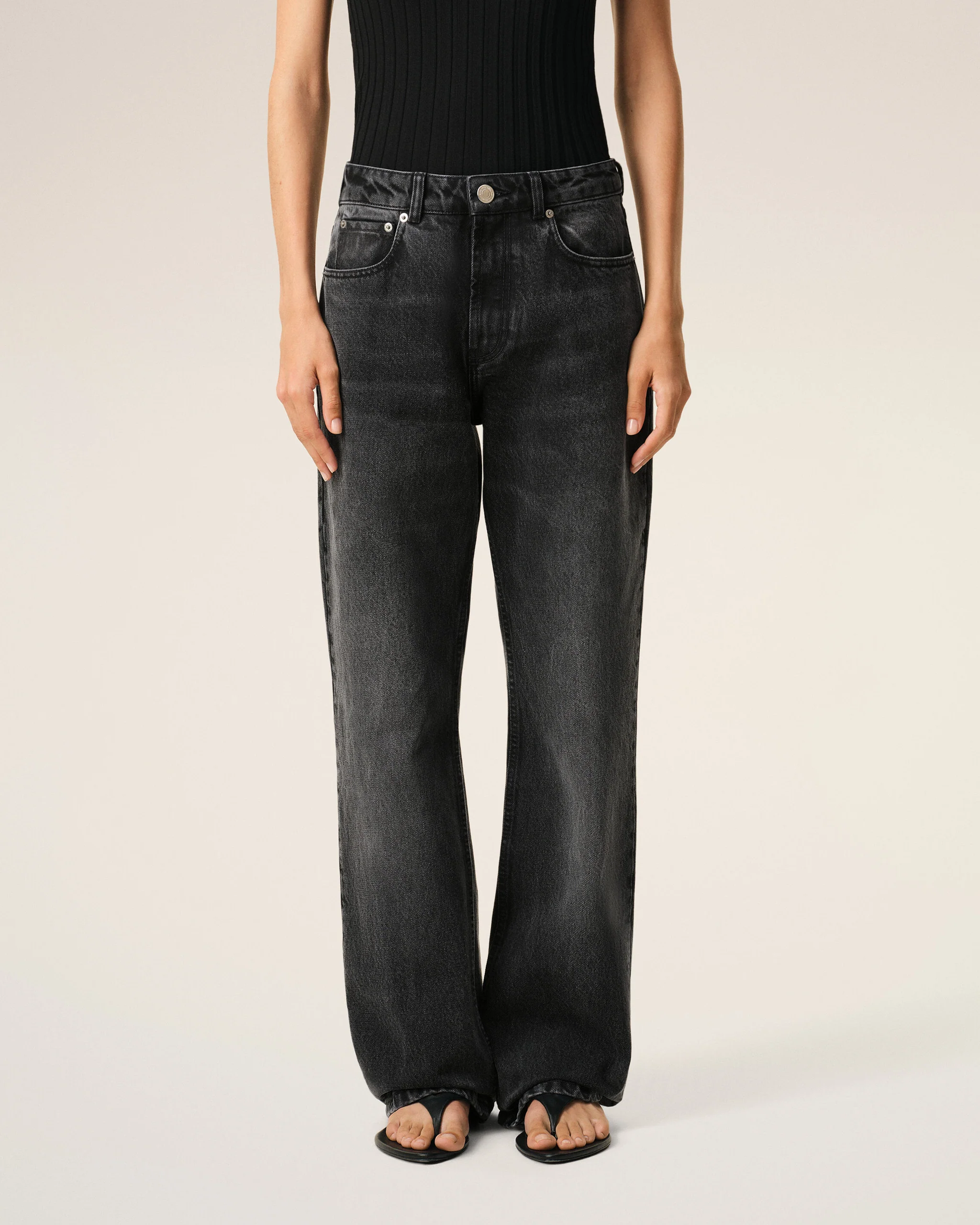 Person from the waist down wearing a black ribbed top tucked into dark washed jeans, standing with open-toe sandals on light background
