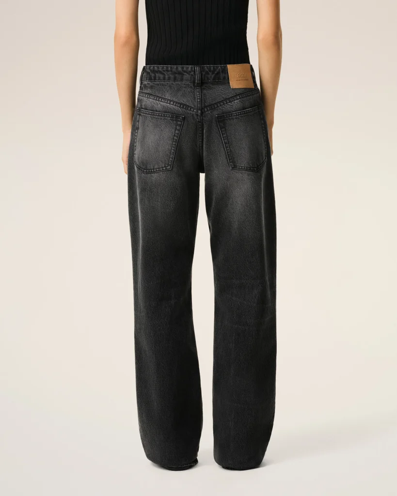 Back view of a person wearing dark, washed denim jeans with large back pockets and a tan leather patch on the waistband progressively visible on the right side.