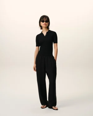 Person wearing a black ribbed polo shirt and wide-leg pants, sunglasses, standing against a pale gradient background.