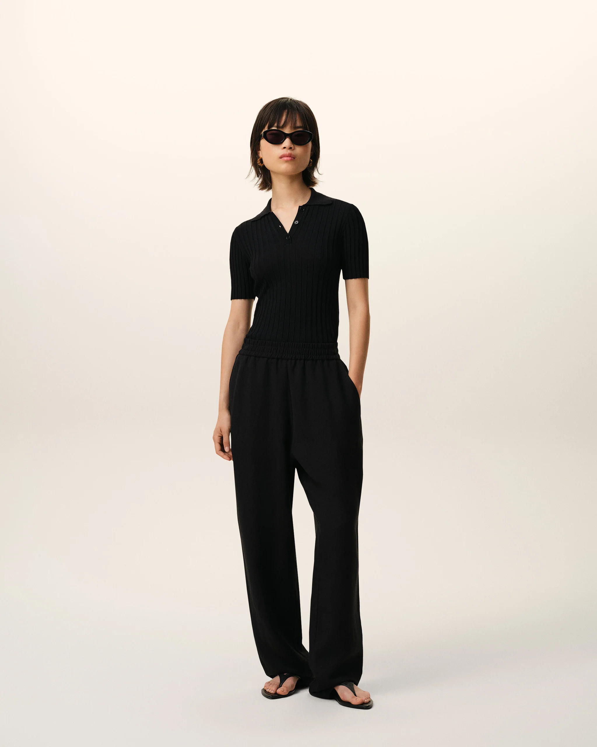 Person wearing a black ribbed polo shirt and wide-leg pants, sunglasses, standing against a pale gradient background.