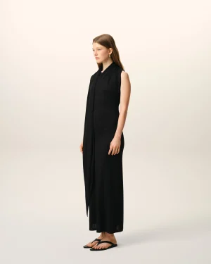 Person standing in profile facing left, wearing a sleeveless black dress and black flip-flops against a pale gradient background.