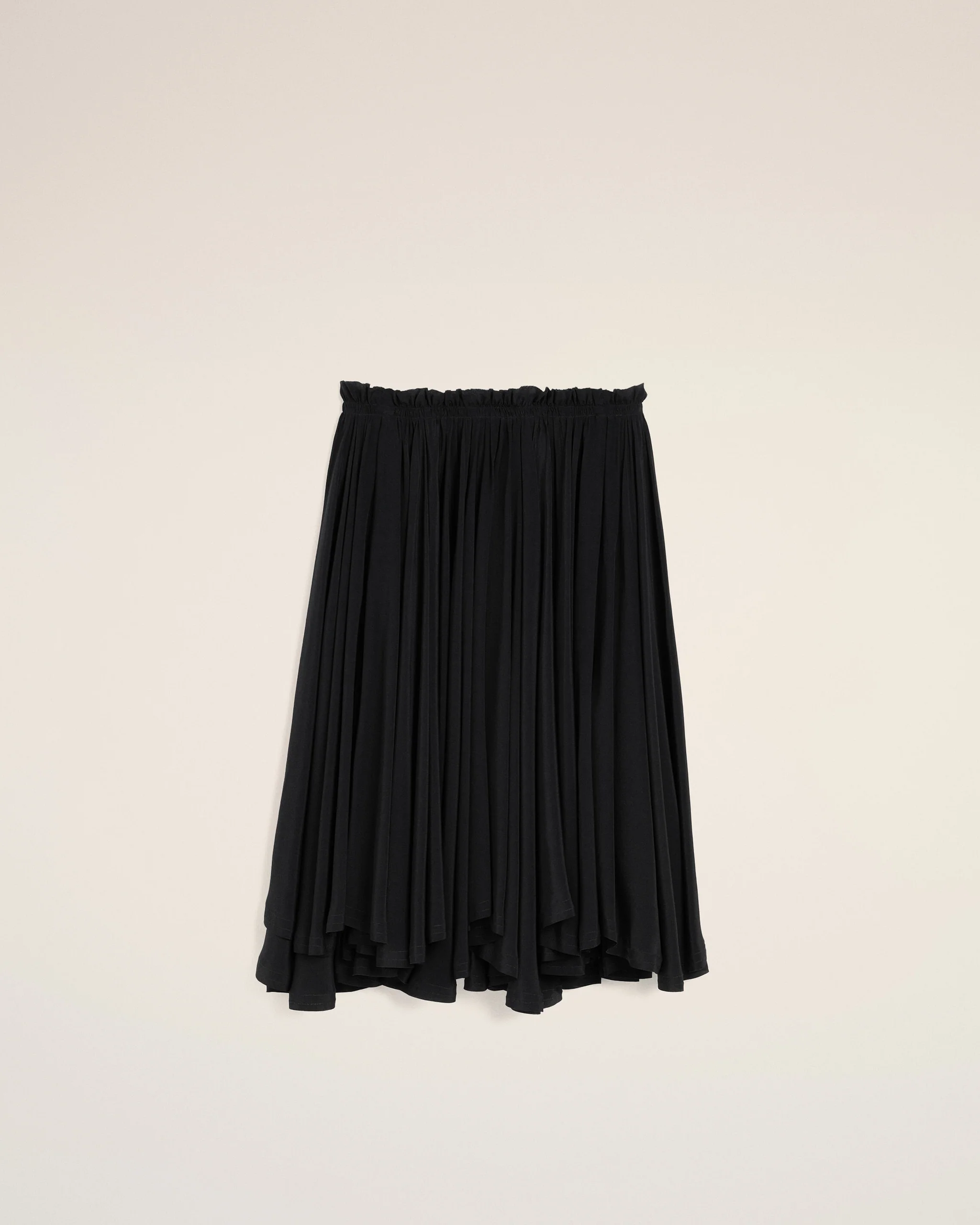 Black pleated skirt hangs against a pale beige wall, displaying its elastic waistband and flowing fabric.