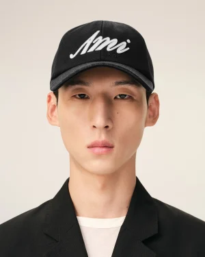 Close-up portrait of a person wearing a black cap with white embroidered script, beige background visible behind.