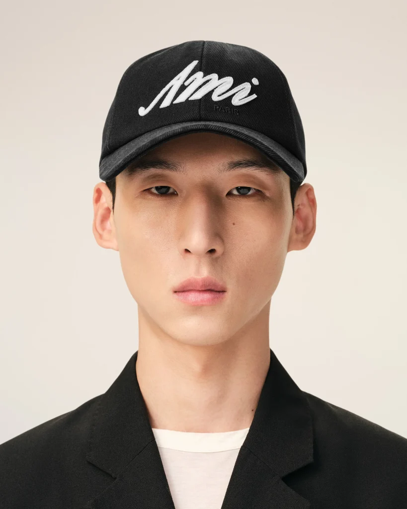 Close-up portrait of a person wearing a black cap with white embroidered script, beige background visible behind.