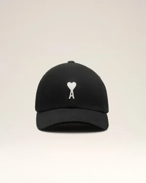 Black baseball cap with a white heart applique on the front panel, against a light beige background.