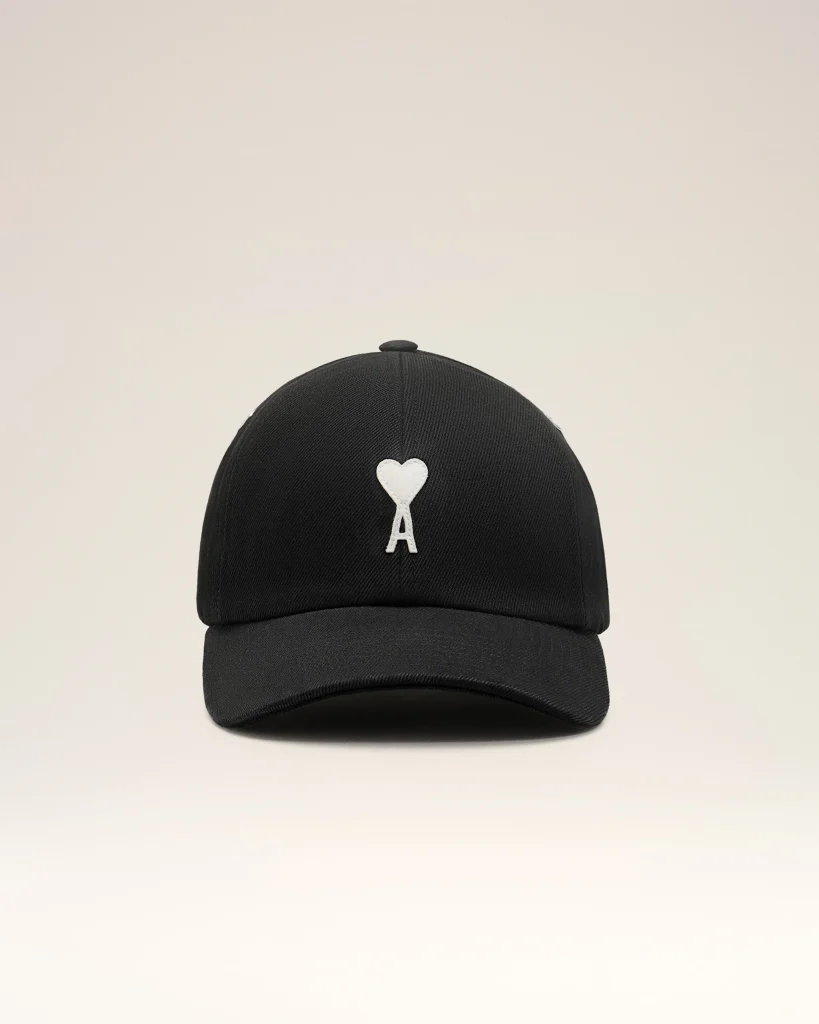 Black baseball cap with a white heart applique on the front panel, against a light beige background.