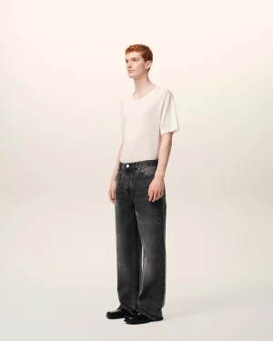 Person standing in a white T-shirt and dark jeans, facing slightly left against a pale gradient backdrop.