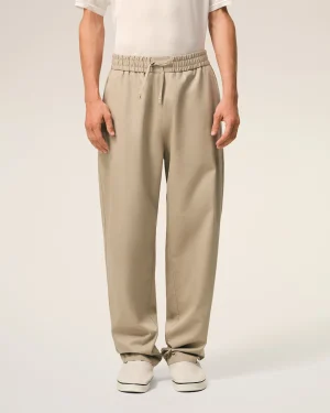 Lower half of a person wearing beige drawstring pants with an elastic waistband and white sneakers against a neutral backdrop.