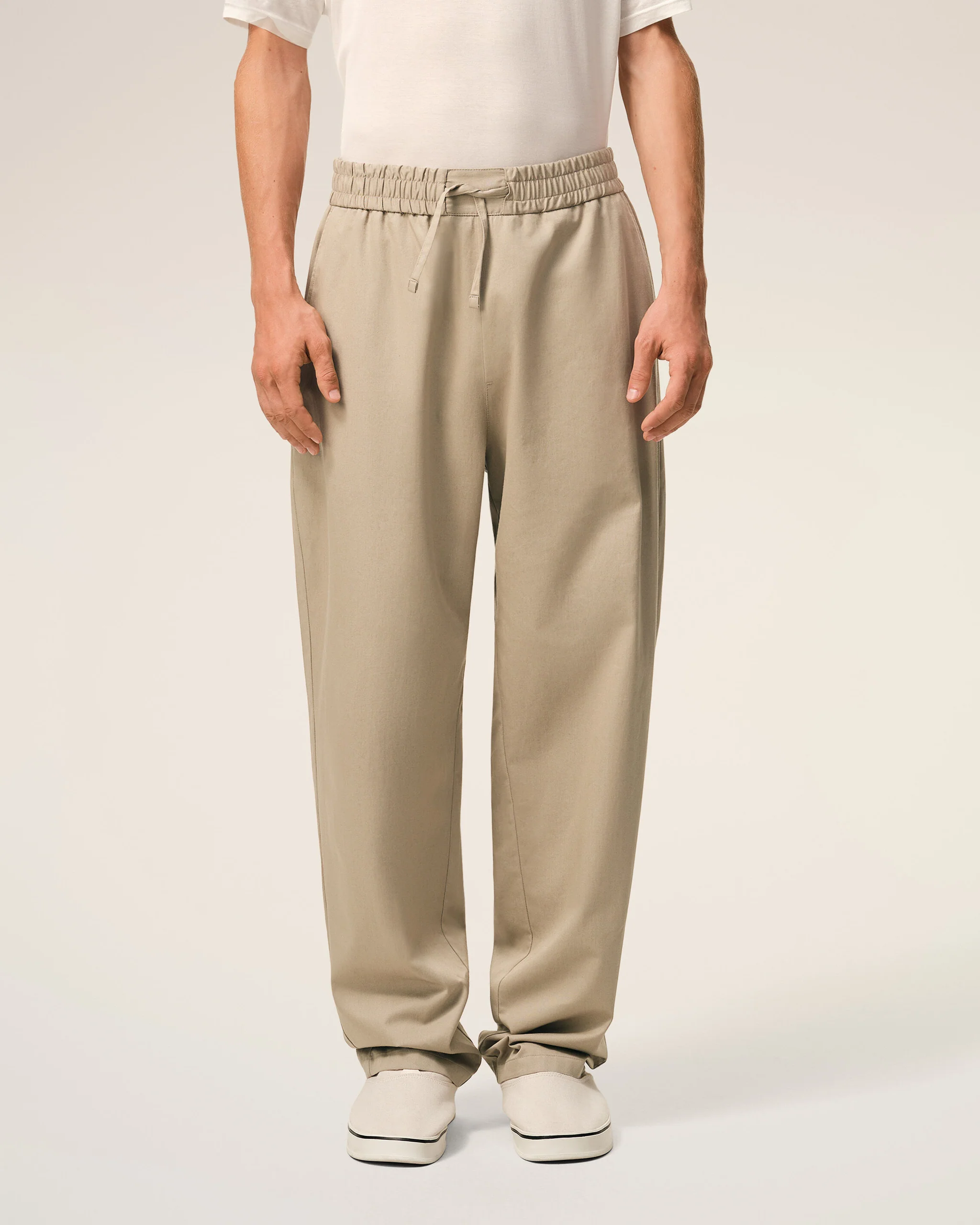 Lower half of a person wearing beige drawstring pants with an elastic waistband and white sneakers against a neutral backdrop.
