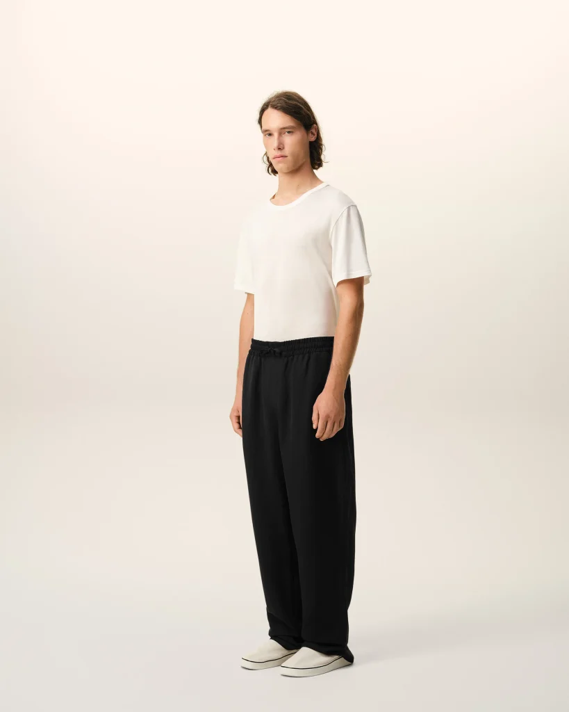 Full-body shot of a person standing, wearing a white T-shirt, black pants, and white sneakers against a pale backdrop.