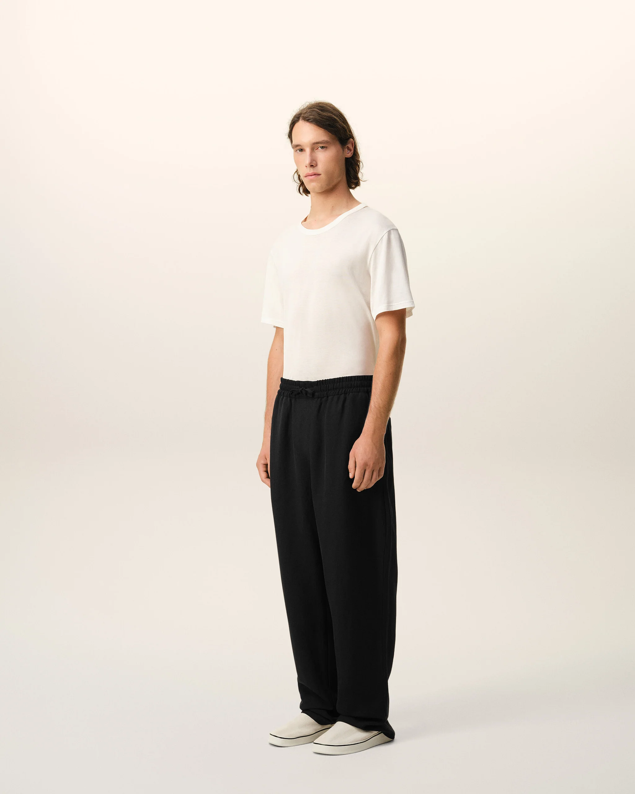 Full-body shot of a person standing, wearing a white T-shirt, black pants, and white sneakers against a pale backdrop.