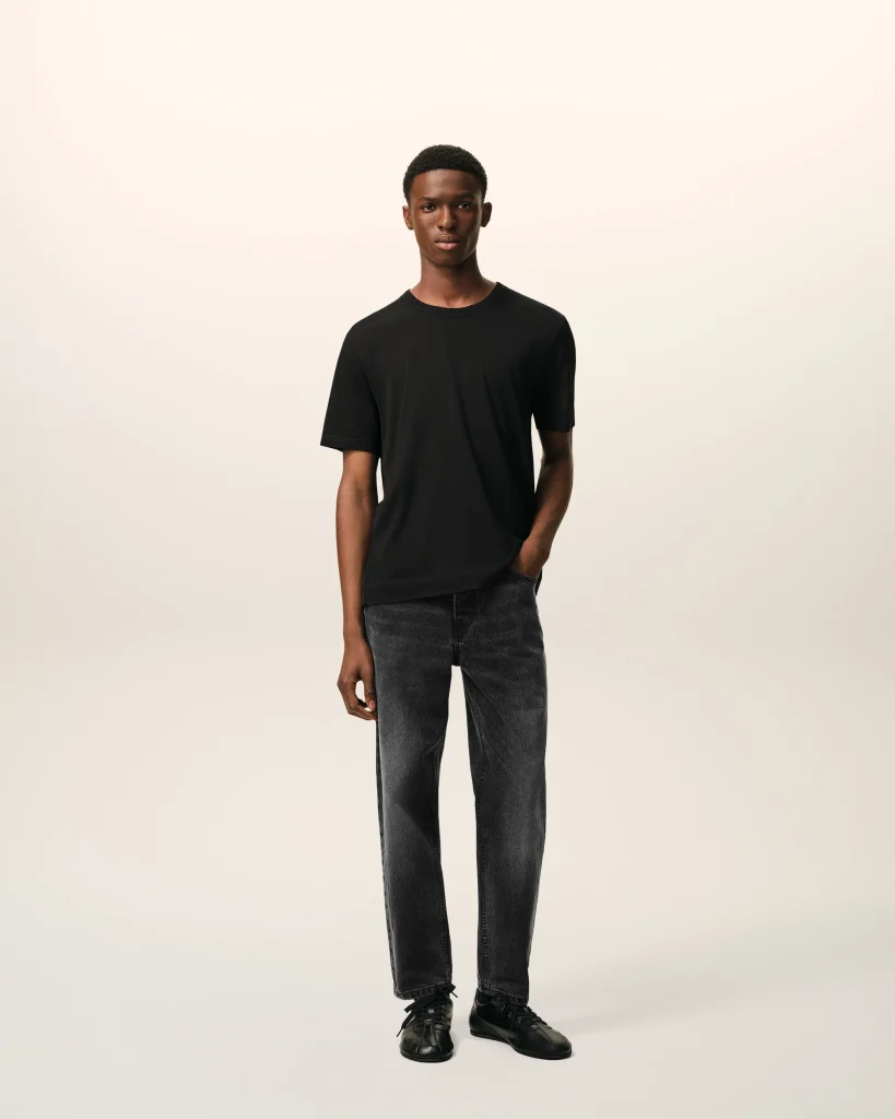 Full-body shot of a person in a black T-shirt, dark jeans, and black sneakers standing against a pale gradient background.