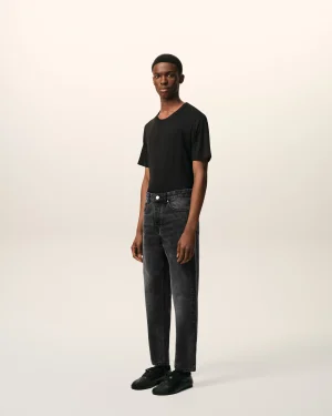 Full-body portrait of a young man in a black T-shirt, dark jeans, and black sneakers standing against a pale gradient backdrop.