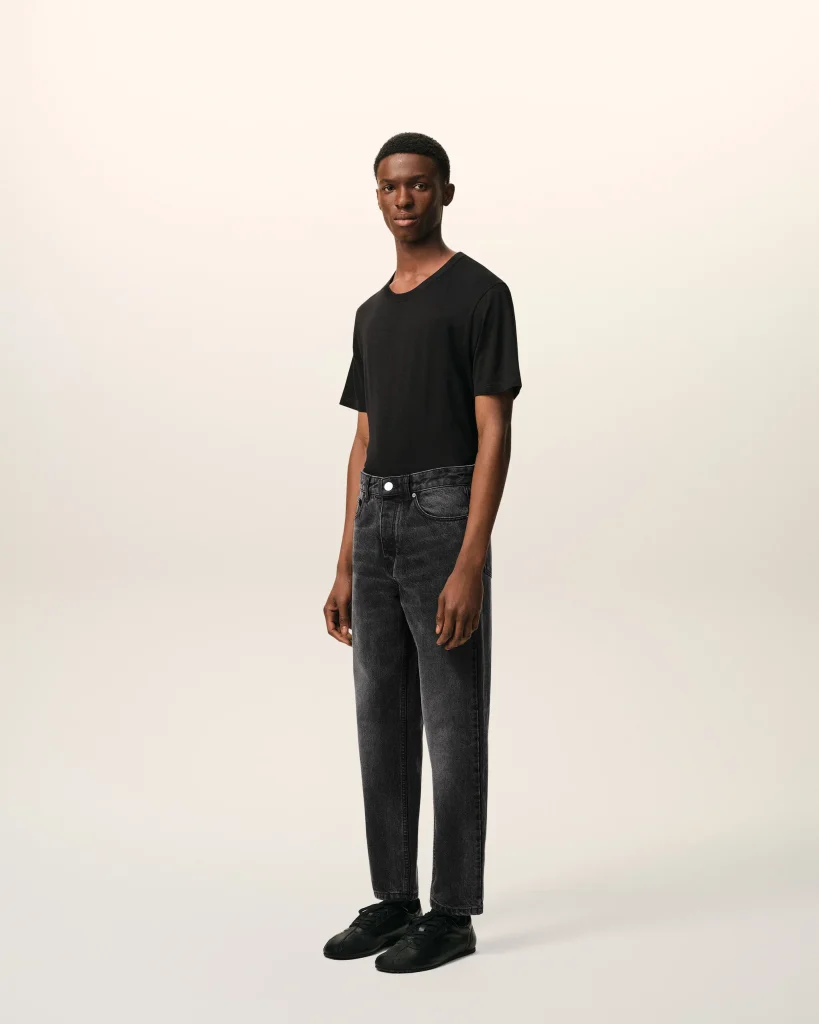 Full-body portrait of a young man in a black T-shirt, dark jeans, and black sneakers standing against a pale gradient backdrop.