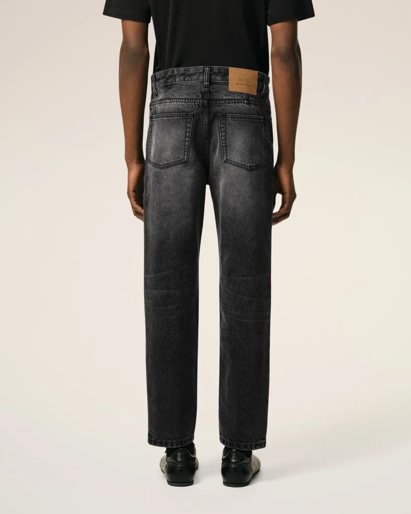 Person standing with back to camera wearing a black T-shirt and dark-washed jeans, leather patch on waistband visible