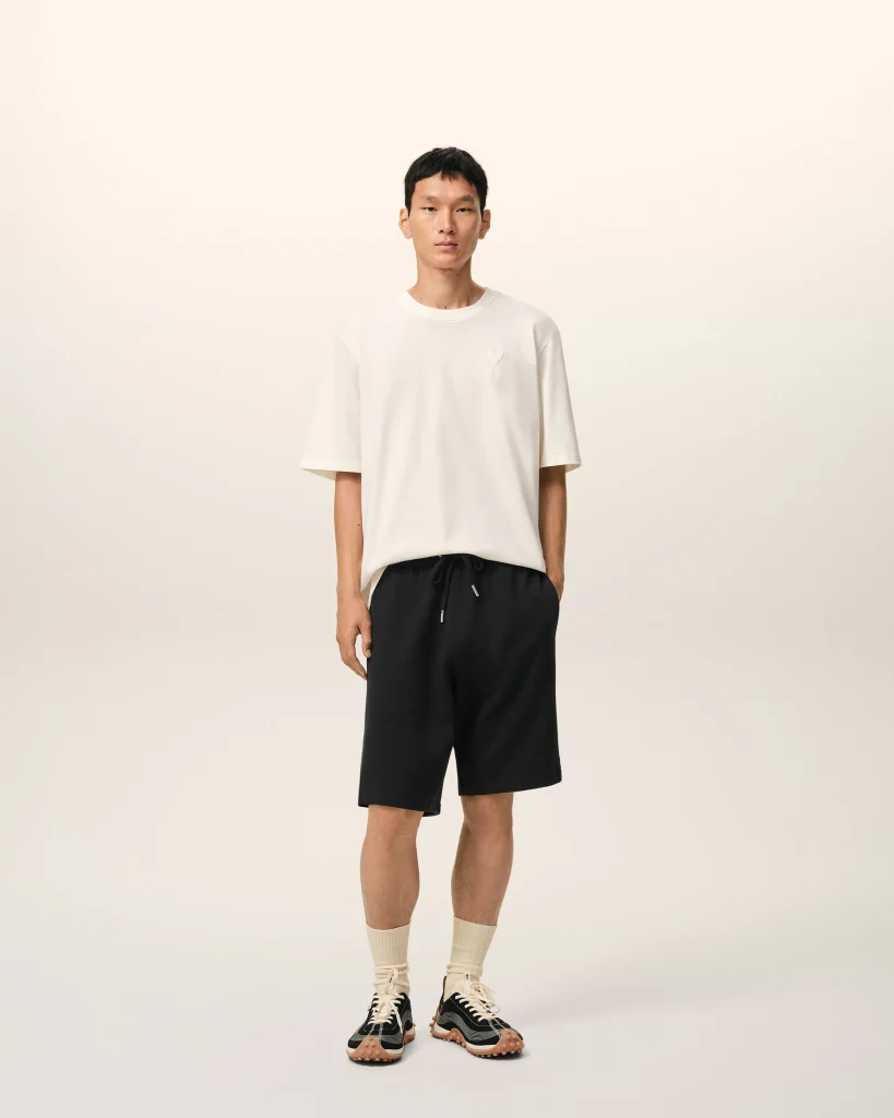 Person standing against a pale background, wearing a white T-shirt, black shorts, beige socks, and black-and-tan sneakers.