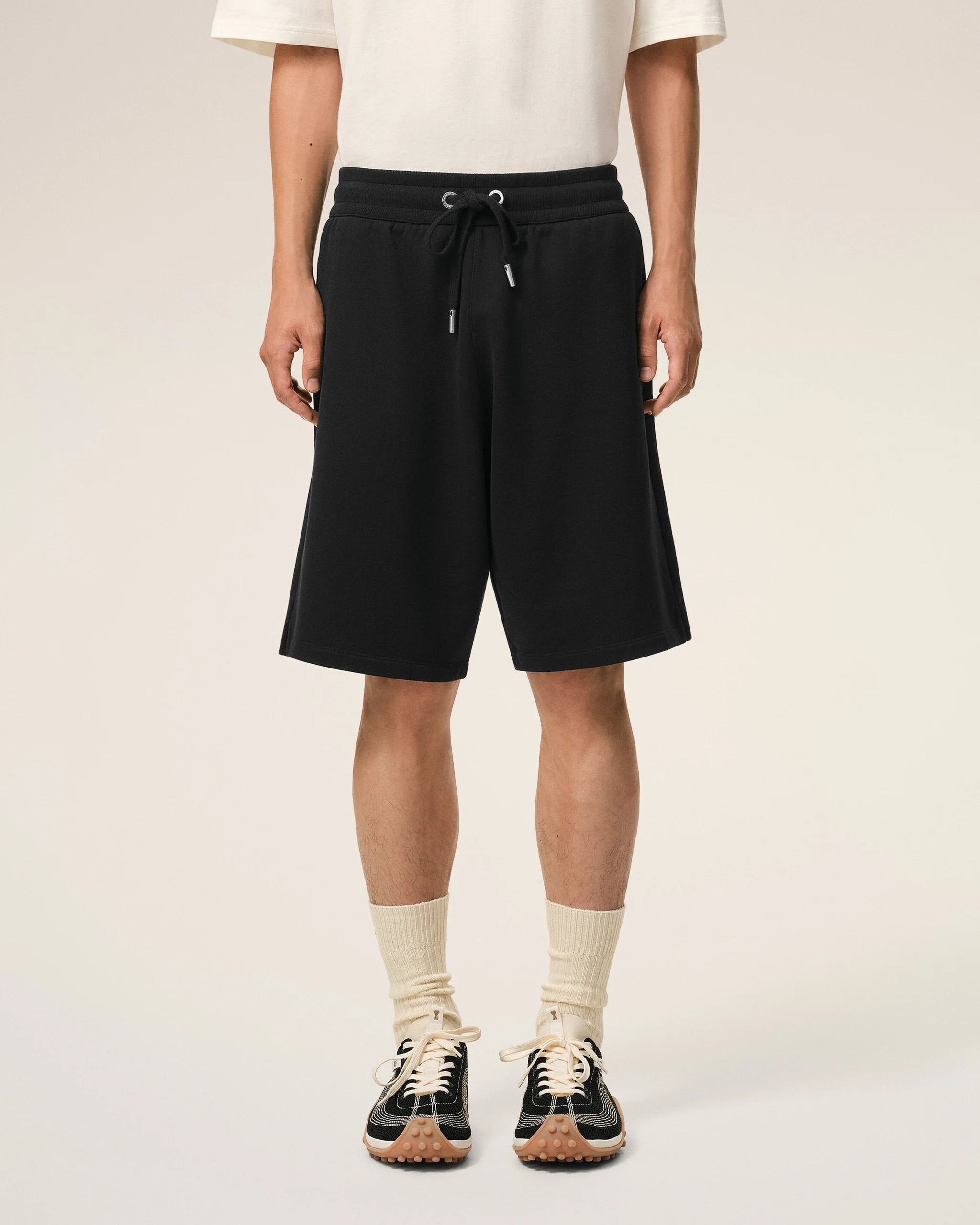 Lower half of a person wearing black drawstring shorts, a white T-shirt, cream socks, and black sneakers, standing against a light background.
