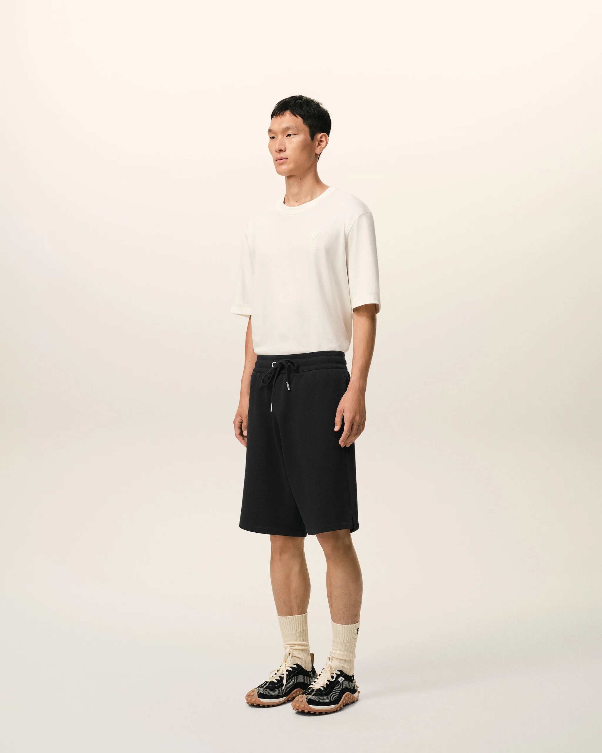 Full-body shot of a man standing facing slightly left, wearing a white T-shirt, black drawstring shorts, beige socks, and black-and-tan sneakers against a light beige background.