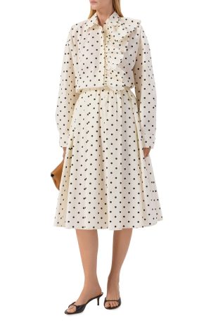 Woman wearing a cream polka-dot midi dress with a ruffled front detail, long sleeves, and black heels, holding a tan clutch.