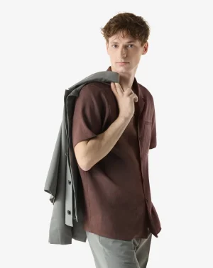 Young man wearing a brown short-sleeve shirt with a gray blazer slung over his shoulder.