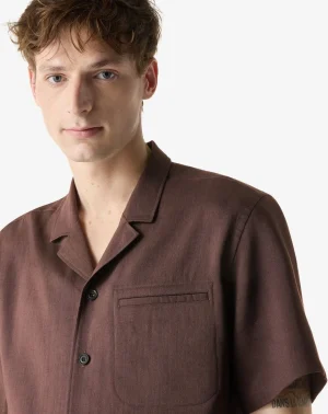 Young man wearing a brown short-sleeve button-up shirt with a chest pocket, facing slightly left against a light background.