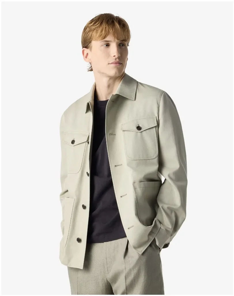 Young man wearing a light beige jacket over a black shirt, with hands in pockets, looking to the right, on a white background.