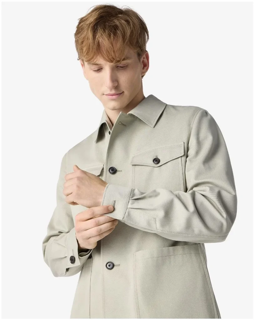 Young man in a light beige trench coat adjusting the cuff on his sleeve.
