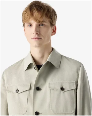 Young man wearing a light beige button-front jacket with large flap pockets and dark buttons.