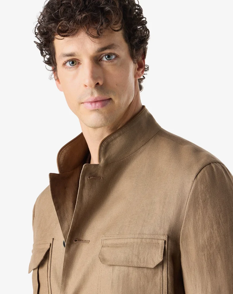 Close-up portrait of a man with curly brown hair wearing a tan coat, looking at the camera against a light background.