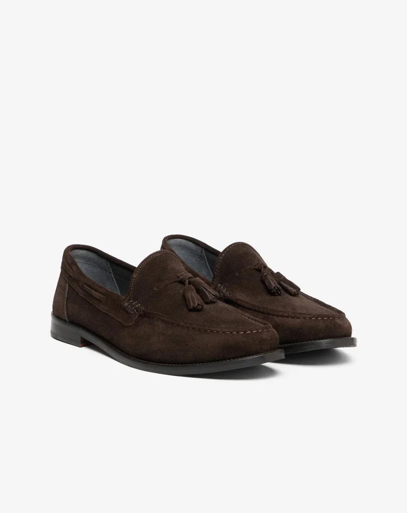 Brown suede loafers photographed from a low angle on a white background, showing the front and side details like tassels and stitching.