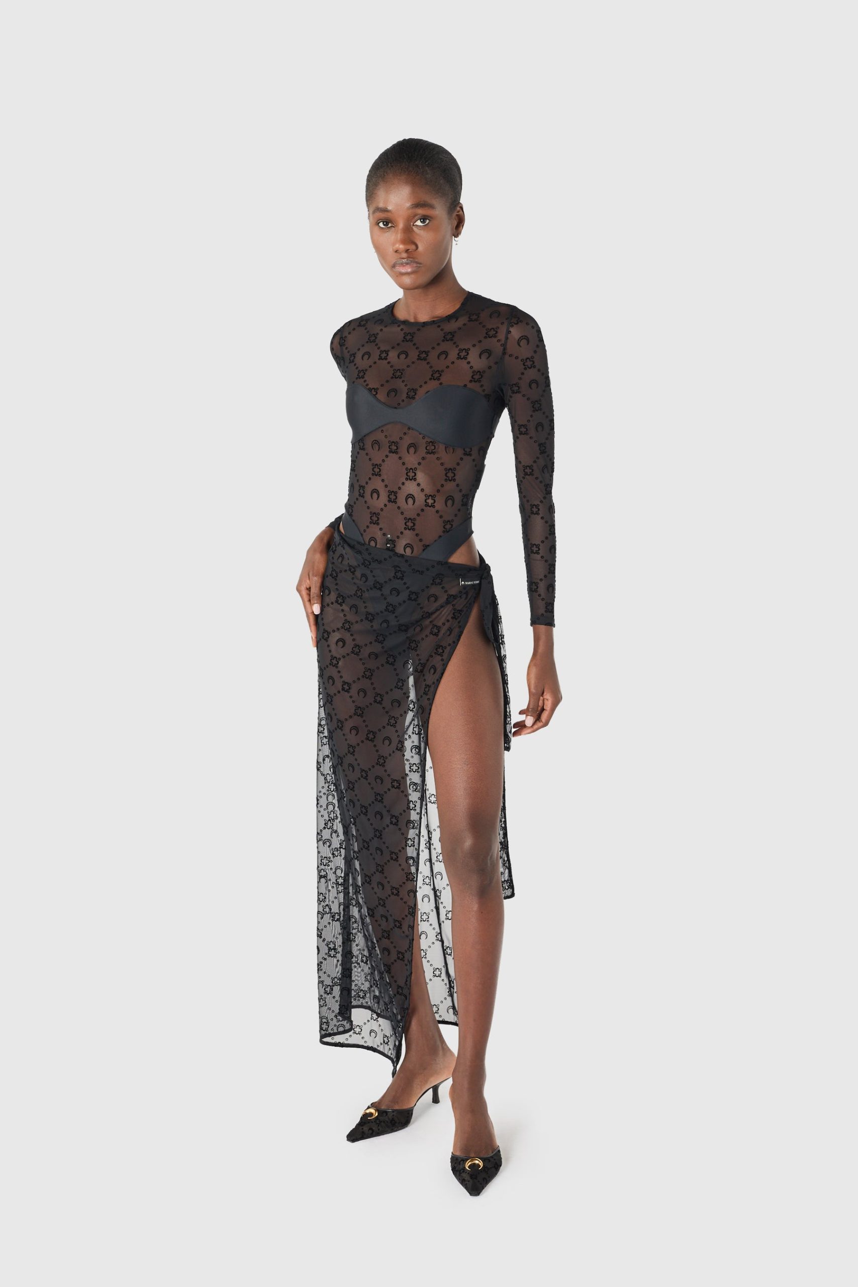 Model wearing a sheer black lace dress with long sleeves and a thigh-high slit, standing against a light background.