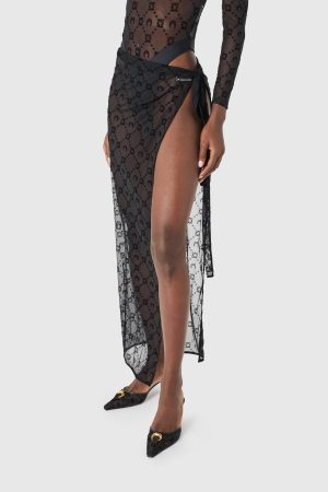 Woman wearing a sheer black lace wrap skirt with a side tie and high slit, paired with black pointed-toe flats with a gold accents bar.