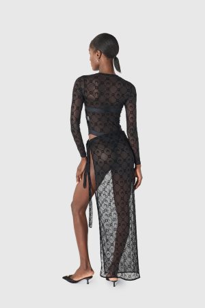 Back view of a woman in a sheer black lace dress with long sleeves and a high slit, wearing black heels.