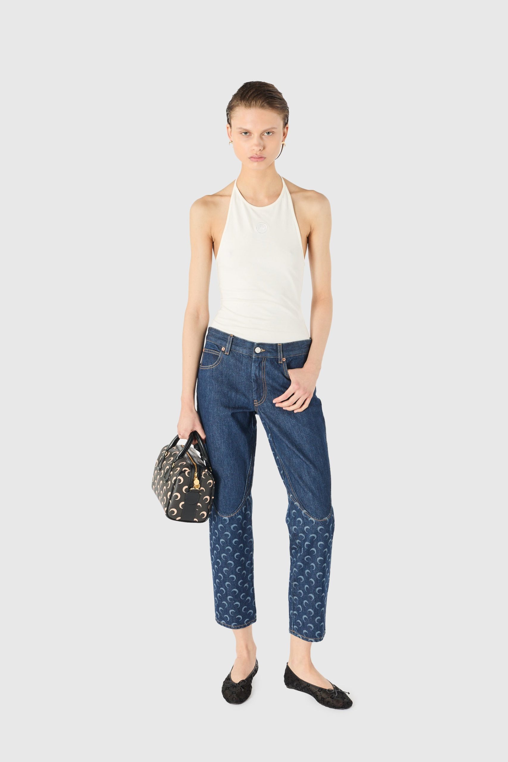 Model standing and facing forward in a white halter top and blue patterned jeans, holding a small patterned handbag.
