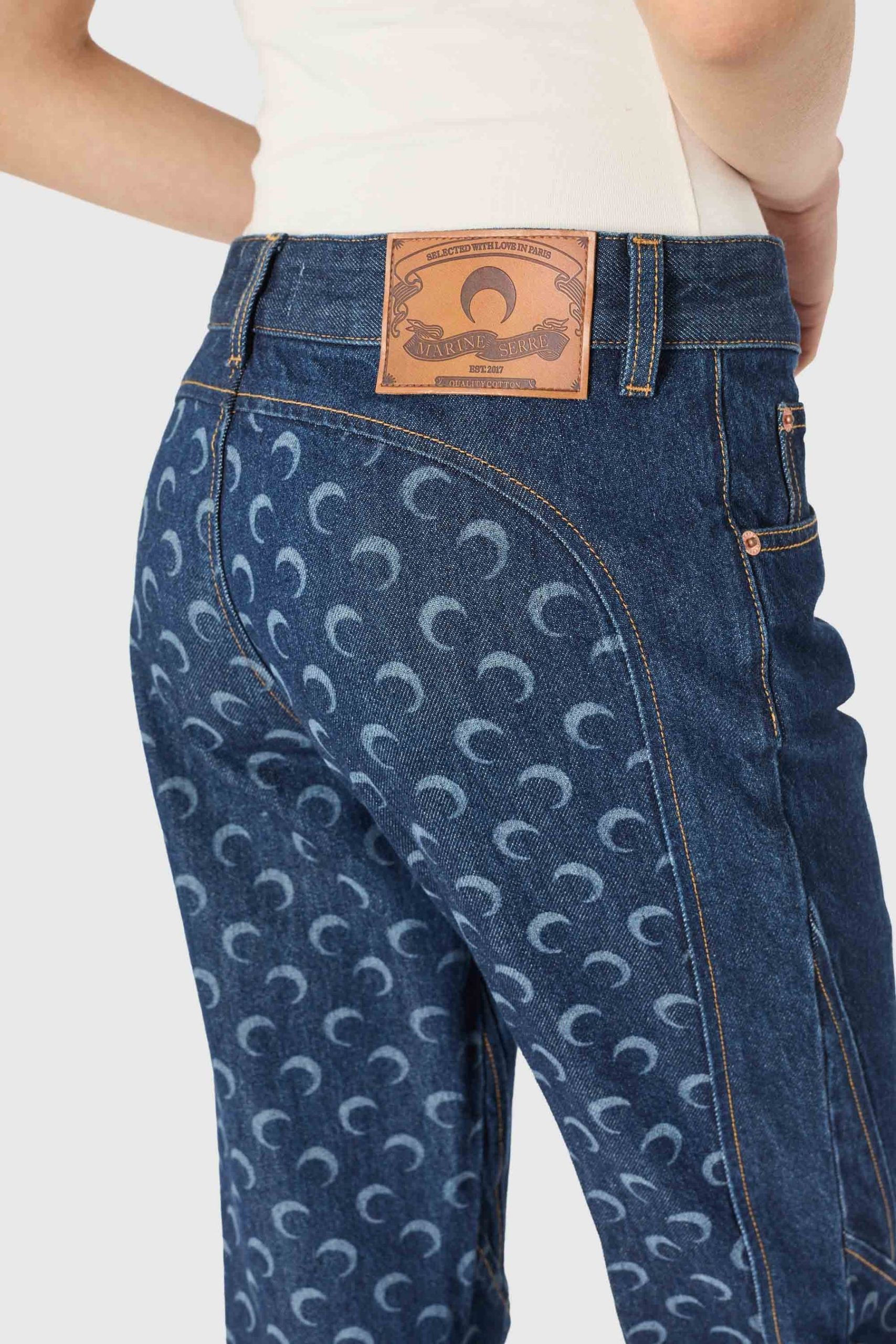 Back view of dark blue denim jeans with a brown leather patch at the waistband and a repeating crescent pattern on the fabric
