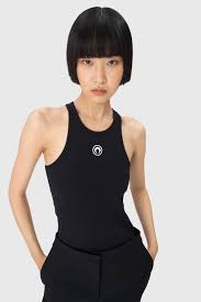 Woman with a short black bob wearing a black sleeveless bodysuit, standing against a light gray background with hands in pockets.