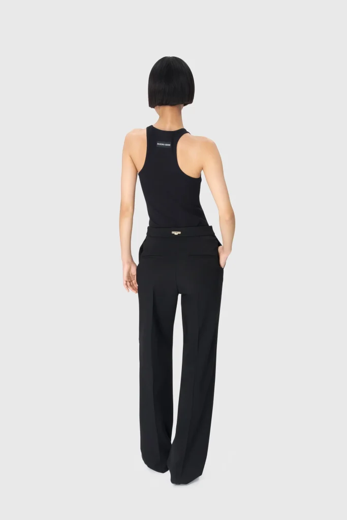 Back view of a person wearing a black sleeveless top and black dress pants, with a short black bob haircut against a light gray backdrop.