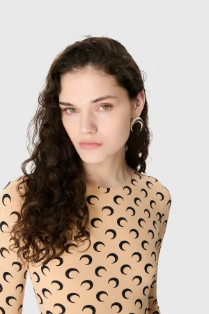 Close-up portrait of a woman with long, curly brown hair wearing a beige garment with black crescent patterns and crescent earrings in front of a light background.