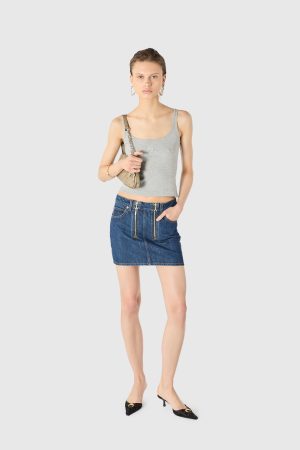 Young woman standing against a light gray background, wearing a gray sleeveless top, a blue denim mini skirt with front pockets, hoop earrings, and carrying a beige shoulder bag.